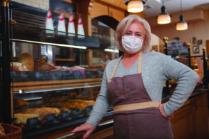 Elderly female baker wearing protective medical face mask working at her shop during coronavirus pandemic