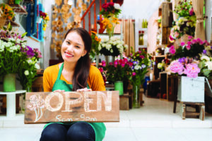 Photo of a happy woman holding an Open sign in front her outdoor flower stand