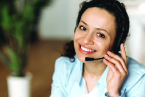 Close-up of mixed race attractive confident adult woman in headset, sitting in office, working as operator of call center or support service, looking directly at the camera and smiling friendly