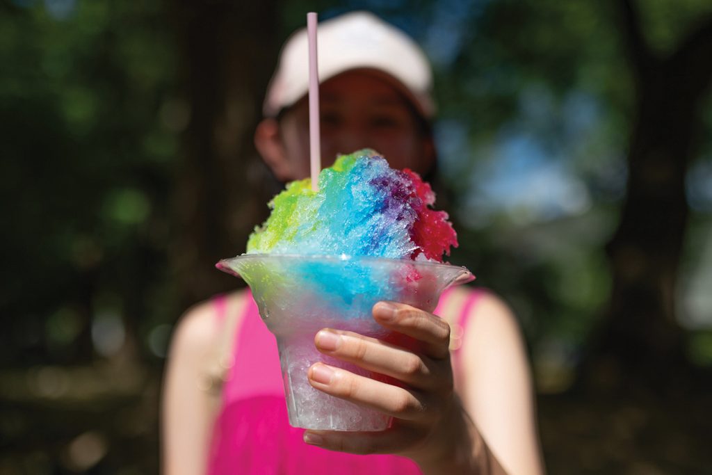 Girl holding out colorful rainbow shaved ice hiding her face